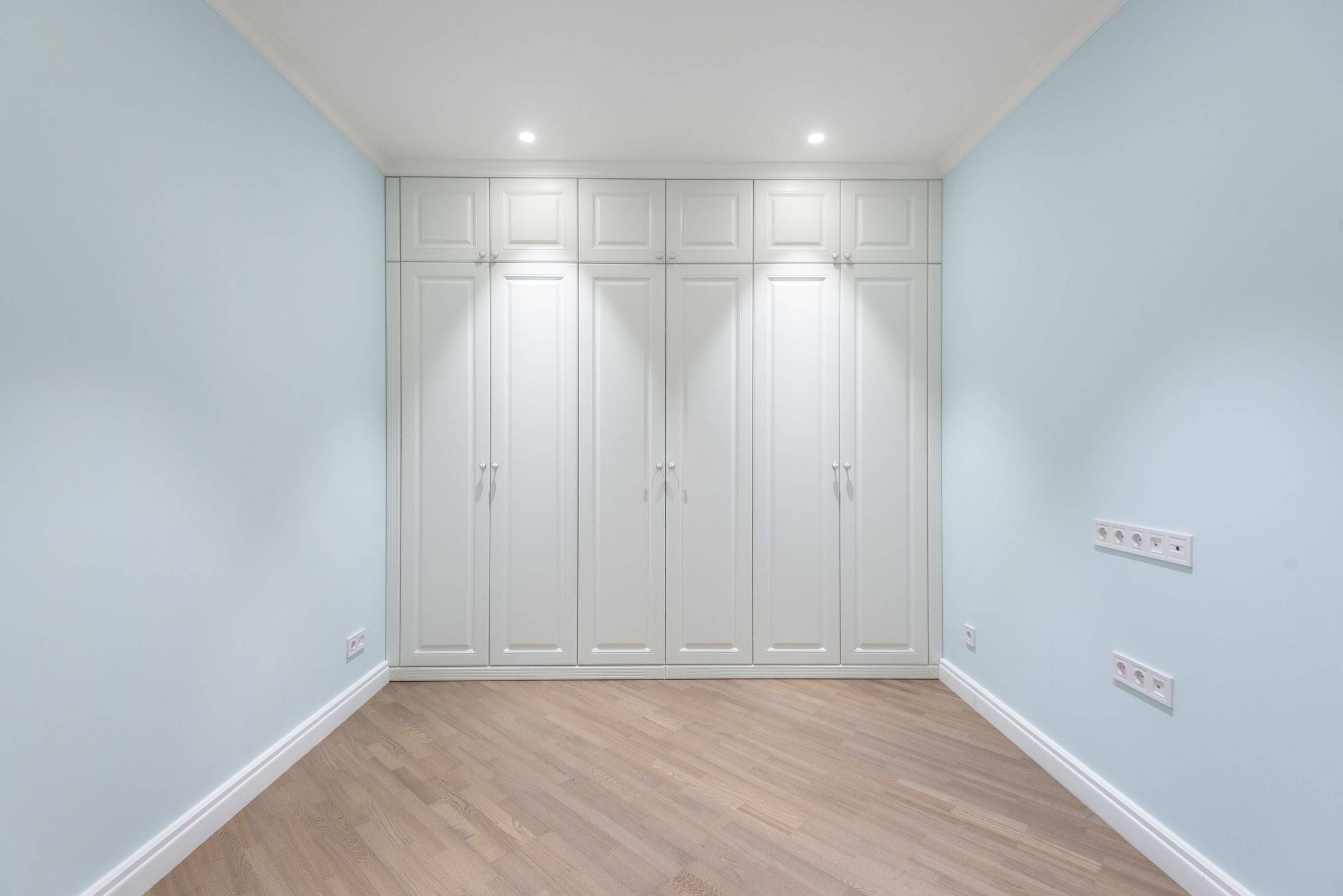 Armoire murale blanche à six portes toute hauteur dans une chambre vide, parquet clair et spots au plafond.