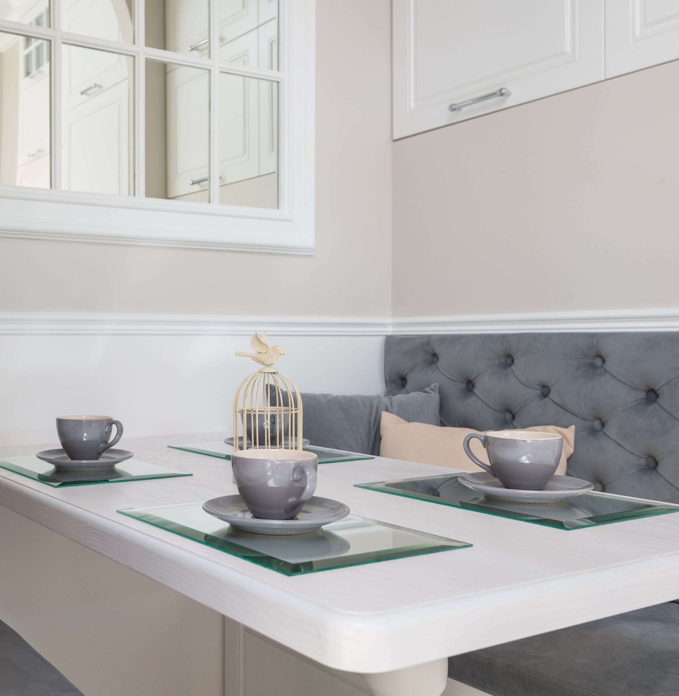 Banquette capitonnée grise contre un lambris blanc, table dressée de tasses.