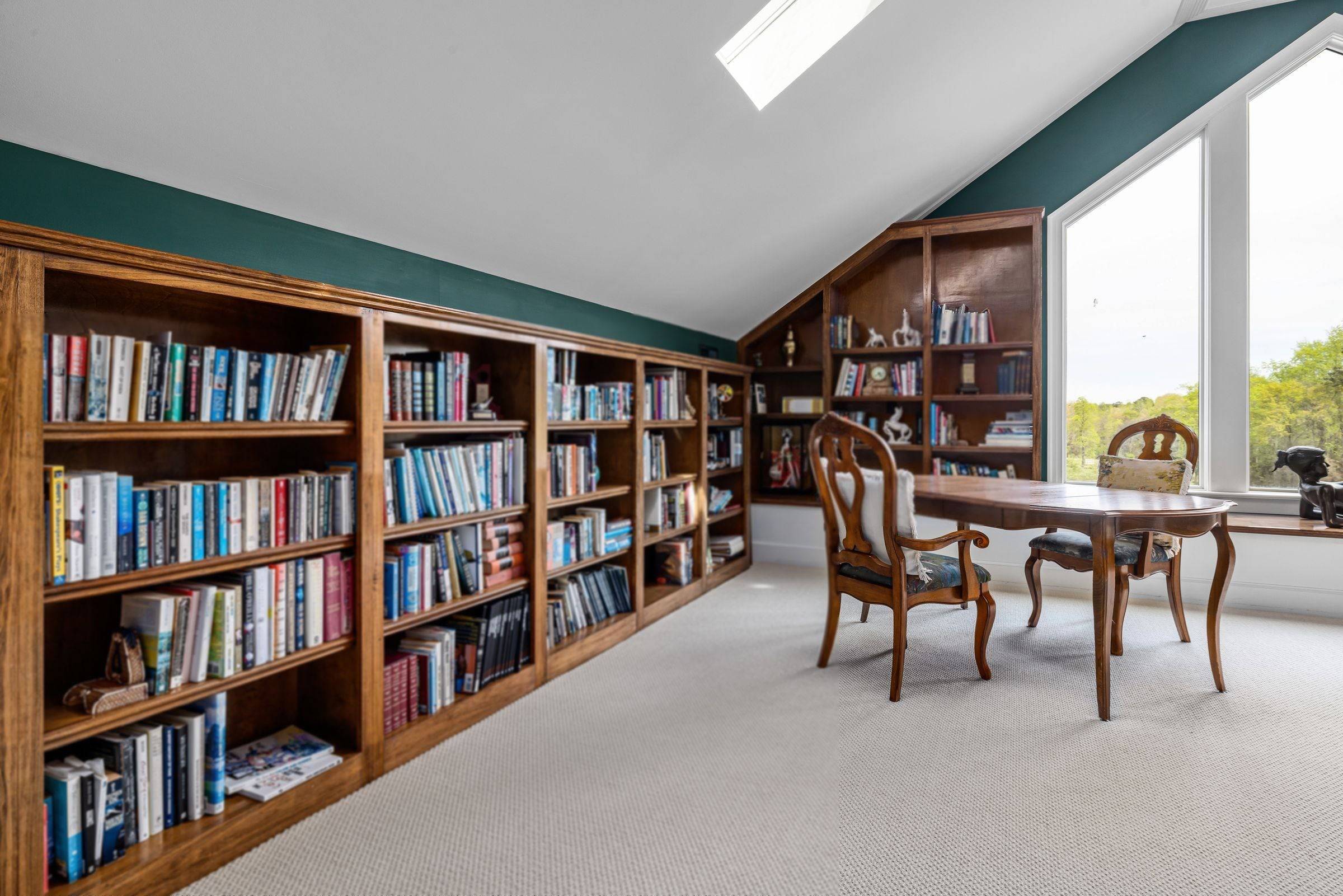 Combles aménagés en salle de lecture, longue bibliothèque en bois sous la pente, table ancienne près d’une grande fenêtre.
