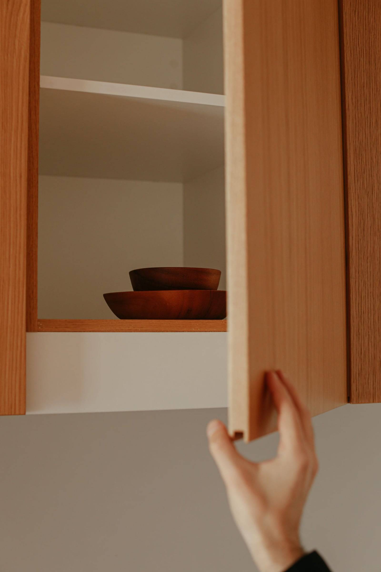 Main qui ouvre une porte d’armoire en bois, tablettes et bols visibles à l’intérieur.