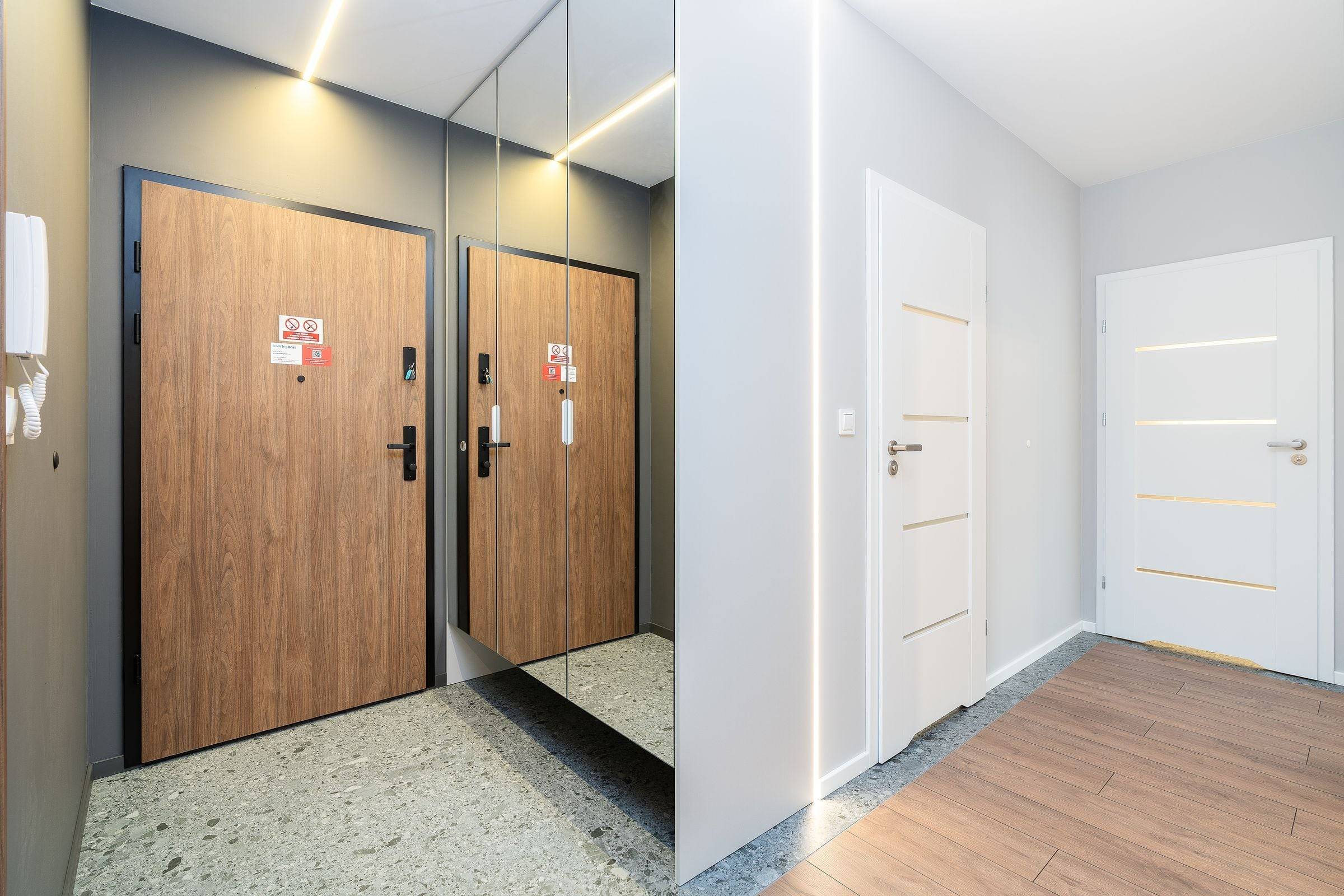Porte palière plaquée bois au cadre noir dans un hall au sol en terrazzo, miroir et porte intérieure blanche.